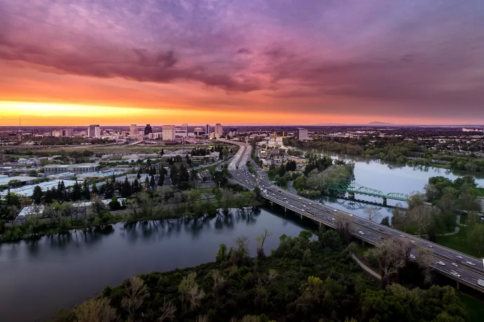 Aerial view of freeway crossing river in Sacramento, California with colorful sunset sky in background.
